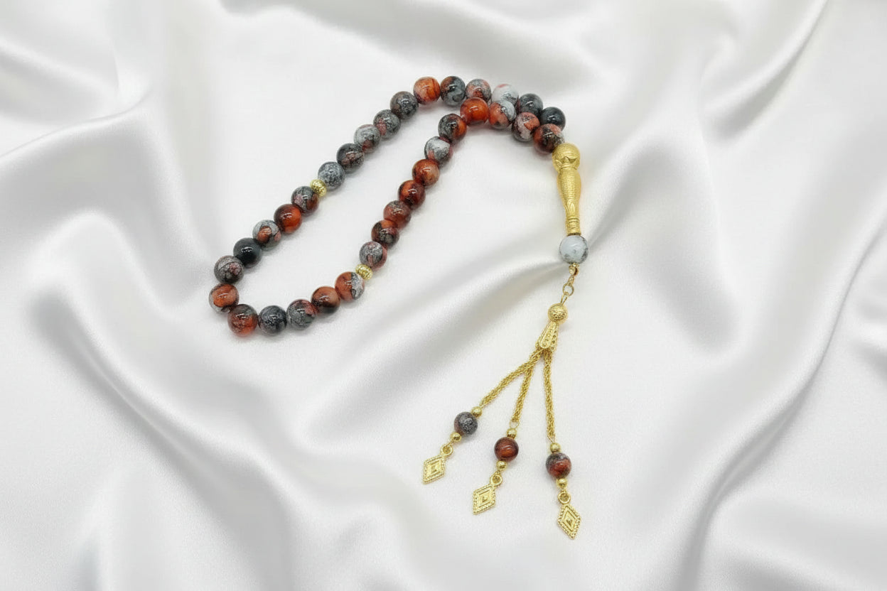 Tasbih de 33 perles de 10mm aux motifs marbrés rouge, gris et noir, avec finitions dorées, séparateurs ciselés et pompon à trois chaînettes dorées sur un drapé de soie blanche.