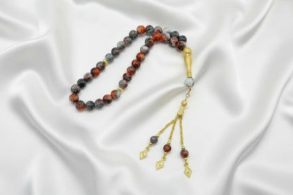 Tasbih de 33 perles de 10mm aux motifs marbrés rouge, gris et noir, avec finitions dorées, séparateurs ciselés et pompon à trois chaînettes dorées sur un drapé de soie blanche.