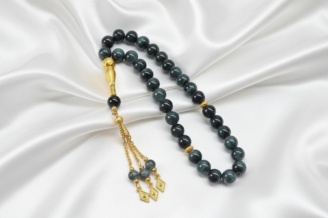 Tasbih de 33 perles de 10mm gris foncé marbré avec des reflets bleu pétrole, séparateurs dorés et pompon artisanal doré à chaînettes.