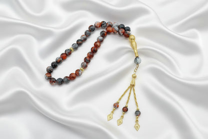 Tasbih de 33 perles de 10mm aux motifs marbrés rouge, gris et noir, avec finitions dorées, séparateurs ciselés et pompon à trois chaînettes dorées sur un drapé de soie blanche.