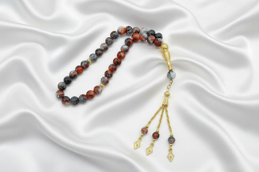 Tasbih de 33 perles de 10mm aux motifs marbrés rouge, gris et noir, avec finitions dorées, séparateurs ciselés et pompon à trois chaînettes dorées sur un drapé de soie blanche.