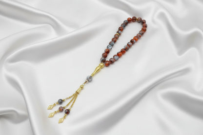 Tasbih de 33 perles de 10mm aux motifs marbrés rouge brique, gris foncé et blanc, avec séparateurs dorés et pompon artisanal luxueux à chaînettes dorées sur un drapé de soie blanche.
