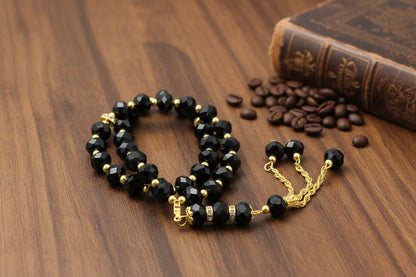 Tasbih élégant avec perles noires facettées et finitions dorées, incluant des anneaux sertis de cristaux et un pompon à chaînettes dorées, sur fond gris neutre.