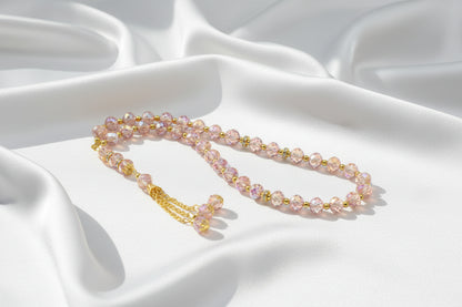 Tasbih de luxe composé de perles roses translucides facettées, avec des anneaux séparateurs dorés incrustés de cristaux et un pompon élégant à chaînettes dorées.