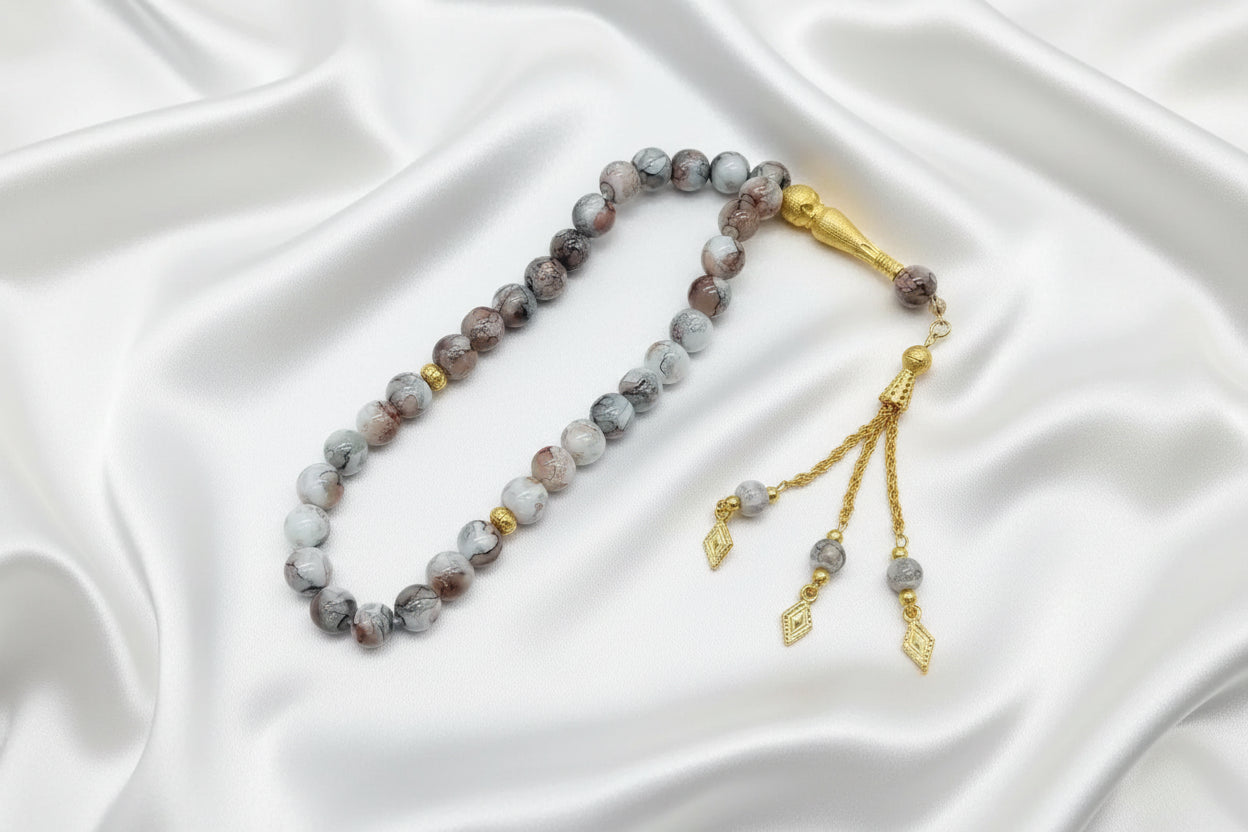 Tasbih de 33 perles de 10mm aux motifs marbrés gris clair, blanc et rose poudré, avec séparateurs dorés et pompon artisanal doré luxueux à trois chaînettes sur fond blanc.