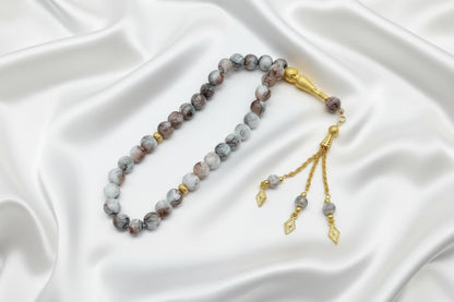 Tasbih de 33 perles de 10mm aux motifs marbrés gris clair, blanc et rose poudré, avec séparateurs dorés et pompon artisanal doré luxueux à trois chaînettes sur fond blanc.