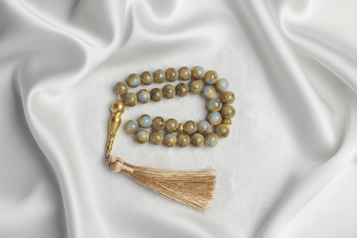 Tasbih de luxe en pierres naturelles aux nuances ocre et sable, orné d'un pompon en fils de soie dorés et éléments en or ciselé, posé sur un satin blanc crème.