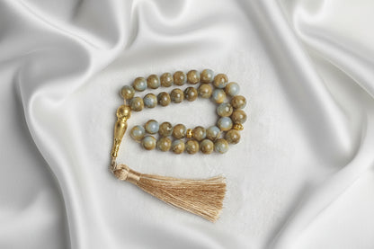 Tasbih de luxe en pierres naturelles aux nuances ocre et sable, orné d'un pompon en fils de soie dorés et éléments en or ciselé, posé sur un satin blanc crème.