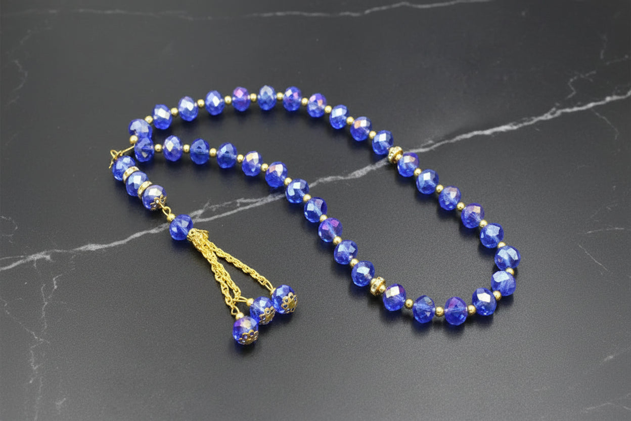 Tasbih élégant composé de 33 perles en cristal bleu saphir facettées, séparateurs dorés sertis de strass et pompon traditionnel à trois chaînettes dorées sur fond sombre.