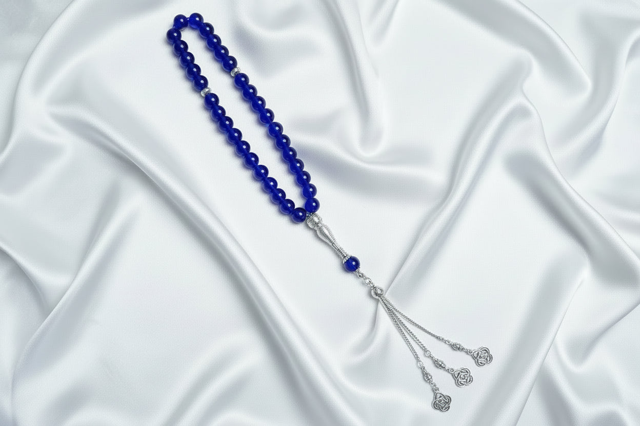Tasbih de 33 perles de 10mm couleur bleu roi intense et brillant, avec séparateurs en argent ciselé et pompon argenté à trois chaînettes terminé par des pendentifs en forme de fleurs délicates, posé sur un drapé de soie blanche.
