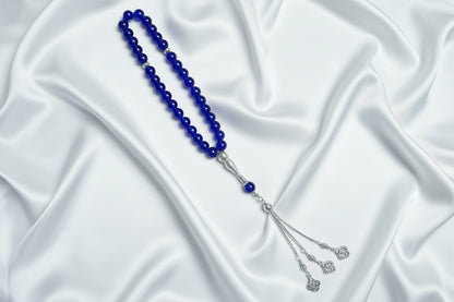Tasbih de 33 perles de 10mm couleur bleu roi intense et brillant, avec séparateurs en argent ciselé et pompon argenté à trois chaînettes terminé par des pendentifs en forme de fleurs délicates, posé sur un drapé de soie blanche.