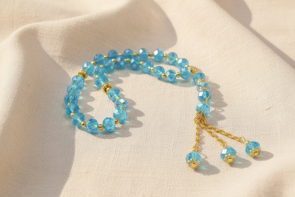 Tasbih élégant en perles de cristal bleu ciel facettées avec reflets irisés, séparateurs dorés sertis de cristaux et long pompon à triple chaînette dorée, posé sur un livre.