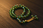 Tasbih circulaire composé de perles vertes irisées et facettées, avec des perles d'espacement dorées, des anneaux sertis de cristaux et un pompon à triple chaînette dorée sur fond gris.